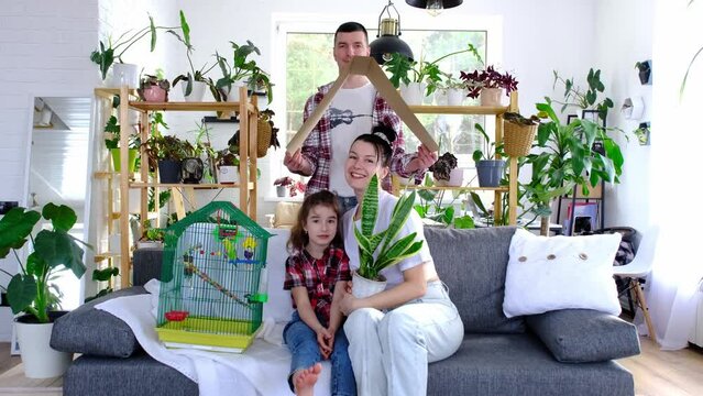 traditional family with a child and a parrot pet in a cage are enjoying a new home, sitting on the sofa and holding the roof. Mortgage, insurance, buying and moving into your own home