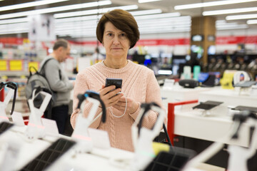 Fototapeta na wymiar European woman who came to an electronics store attentive selects a mobile phone near a shelf with goods to buy them