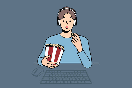 Man With Package Of Popcorn Watches Movie Sitting At Table With Computer And Looks At Screen Intrigued. Funny Guy In Headphones Is Relaxing Watching New Movie Blockbuster Or Popular Tv Series