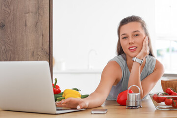 Young woman with laptop and vegetables cooking in kitchen