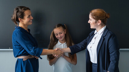 The teacher shakes hands with the mother of the schoolgirl standing at the blackboard. 