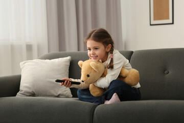 Little girl changing TV channels with remote control on sofa at home