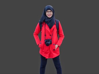 Portrait of smiling young Muslim woman wearing black hijab and red jacket with DSLR camera hanging from neck hands in pockets. Happy and cheerful expression. 