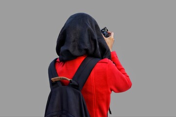 Close up portrait of young Muslim woman wearing black hijab and red jacket with backpack looking through viewfinder and taking picture with DSLR camera.