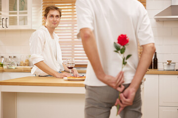 young gay couple looking at boyfriend and holding rose behind his back for surprise in the kitchen