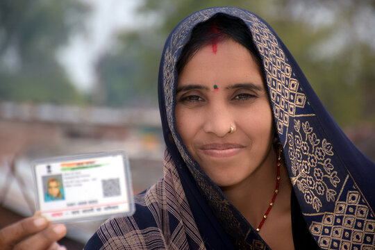 Indian rural woman wearing a sari with smiling face shows her blurred aadhar card in her hand, blurred rural background.