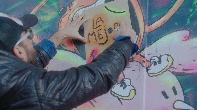 Graffiti artist using paper stencil and spray paint while adding a sign La Mejor (The Best translation in English) to artwork on the wall outdoors. Handheld camera medium shot