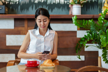 Beautiful Asian woman sitting and playing with smartphone in a coffee shop. While waiting for the appointed friends to sit and talk, there was coffee and croissants on the table.