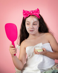 Playful young girl looking in mirror after putting on facial cleansing mask - 630950029