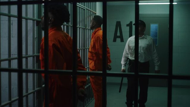 Two prisoners in orange uniforms stand facing metal bars in front of prison cells. Jailer with baton walks behind criminals and talks with them. Murderers serve imprisonment term in detention center.