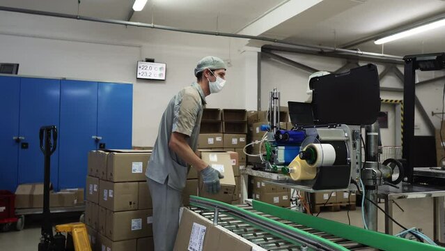 Factory Man Takes a Packed Cardboard Box From the Packaging Line And Puts It In a Stack For Storage. Packaging Line At Modern Factory. Production Prepared For Storage. Modern Packaging Line process