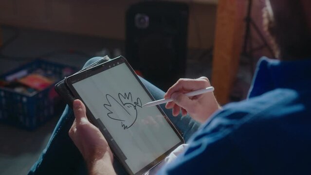 Professional illustrator drawing cartoon style bird sketch on a touchscreen of a digital tablet, using stylus and eraser tool on device. Over the shoulder view, close-up shot