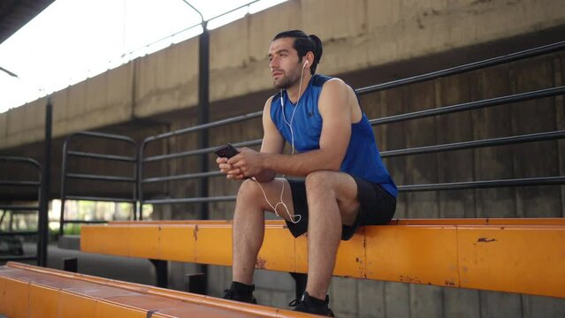 Caucasian sportsman basketball player sitting on bench and listening to the music on headphones at outdoors court under highway in the city waiting for friends playing streetball together in sunny day