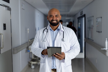Portrait of happy biracial male doctor wearing lab coat holding tablet in corridor at hospital - Powered by Adobe