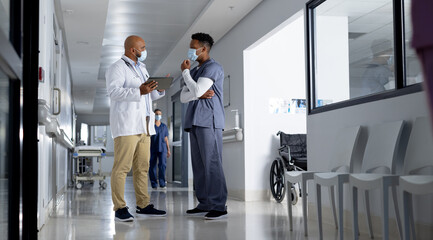 Diverse male doctors wearing face masks using tablet and talking in corridor at hospital