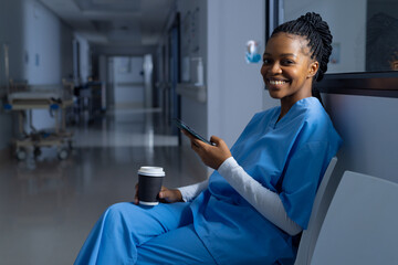 Portrait of african american female doctor wearing scrubs using smartphone in corridor at hospital