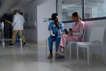 Fototapeta premium Diverse female doctors wearing scrubs drinking coffee and using smartphone in corridor at hospital