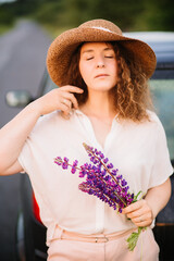 Young woman stands in white shirt near car with purple and pink lupins. Beautiful young woman in hat with curly hair with bouquet of lupins. Sunset or sunrise, bright evening light
