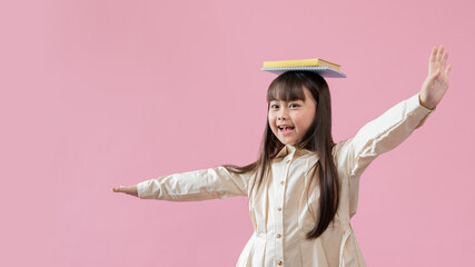 A cheerful Asian girl puts books on her head and makes a flying gesture, isolated pink background