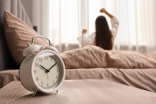 It's lazy morning o'clock. Alarm clock on bedside table and woman stretching in room, selective focus
