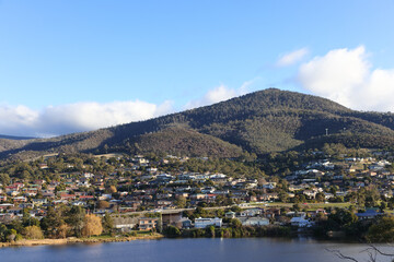 Fototapeta na wymiar Residential houses nestled at the bottom of a large mountain with a cloudy blue sky on the Derwent River in Tasmania 
