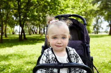 Baby in stroller on a walk in summer park. Adorable little girl sitting in pushchair, funny facial expression. Child in buggy