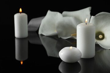 Burning candles and white calla lily flowers on black mirror surface in darkness, closeup with space for text. Funeral symbols