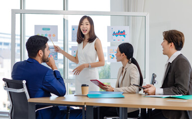 Millennial Asian professional successful businesswoman lecturer presenter in formal wears standing using pencil pointing presenting infographic paperworks documents on glass board in meeting room