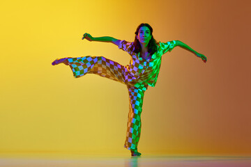 Young artistic, talented woman dancing contemporary, freestyle dance against gradient yellow orange background in neon light. Concept of modern dance style, hobby, art, performance, lifestyle, ad