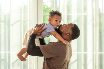 Happy african black parents dad father throw baby son on piggyback and neck riding teasing fun near window. Black baby son and daddy enjoy teasing kiss cheek and neck riding at window light