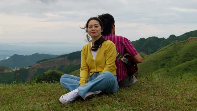 A couple sitting back to back in the mountains, Man playing guitar sitting on the grass surrounded by mountains, Slow motion