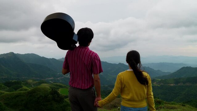 A couple holding hands walking in the mountains, Men holding guitar on his shoulder walking in the scenic outdoors with his girlfriend, Slow motion