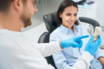 Male dentist explaining to smiling woman patient way of treatment on plastic model
