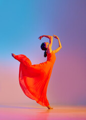 Elegant, graceful young woman dancing in red dress against gradient multicolored background in neon...