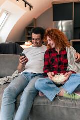 An interracial couple is sitting on the couch and laughing. She sits with popcorn in a bowl on her lap, he sits next to her with his arm around her and a phone is in his other hand
