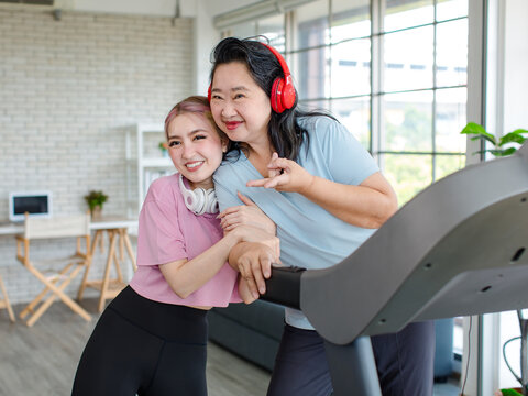 Asian happy healthy old chubby retirement pensioner mother smiling standing posing taking photo with young daughter wear headphones showing thumb up exercise on adjustable running pad in living room