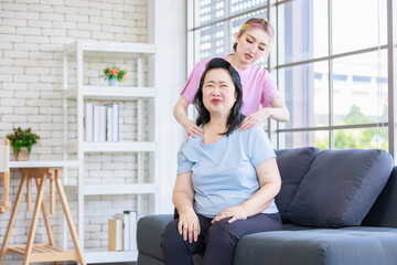 Asian young beautiful daughter standing behind massaging neck of old senior pensioner having backache from stretching in casual sportswear sitting on sofa couch in living room at home.
