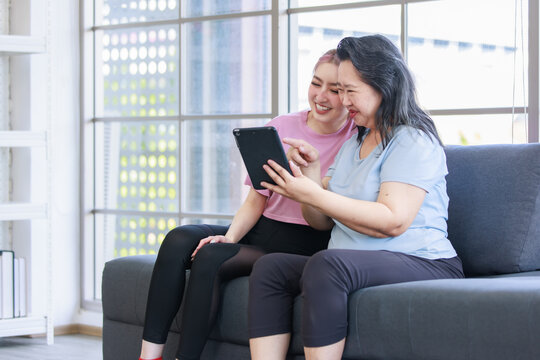 Asian young beautiful daughter and old senior pensioner retirement cheerful happy healthy mother in casual sportswear sitting on sofa couch in living room at home using tablet video call with family