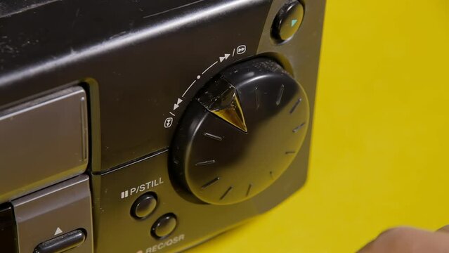 Angled close-up shot: a hand manipulates the knob of a vintage VHS VCR, commanding the videotape motor to perform various actions such as fast forward and rewind.