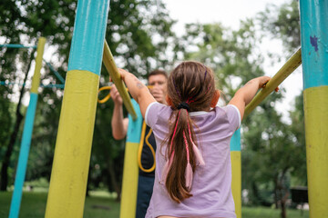 Father with child girl doing pull-ups on workout outdoor area. Healthy active lifestyle, happy family time. Modern fatherhood concept 