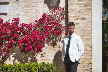 Attractive young businessman with beard, suit and tie, hands in pockets, posing next to a wall with a background of flowers. Concept beauty, fashion, success, achiever, trend.