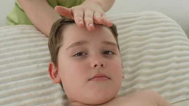 Closeup face of five year old boy receiving osteopathic or chiropractic treatment in pediatric clinic, looking at camera. Close up of unrecognizable female osteopath doing physiological skull therapy.