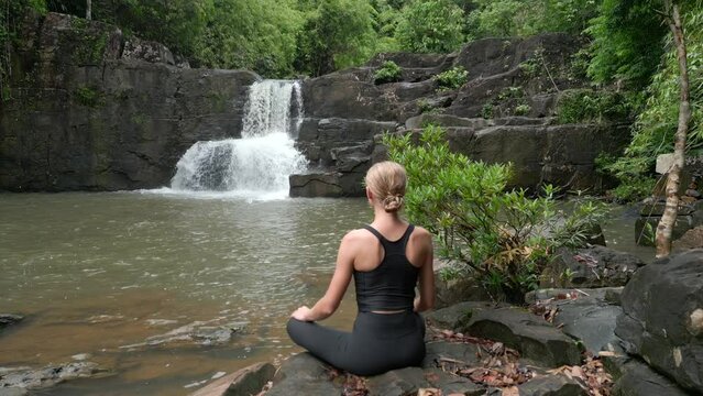 Beautiful young woman practice meditation at an amazing waterfall in the tropical rainforest in Thailand.