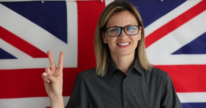 Teacher with glasses gives Victory sign gesture honoring language learning. English tutor stands in front of British flag slow motion