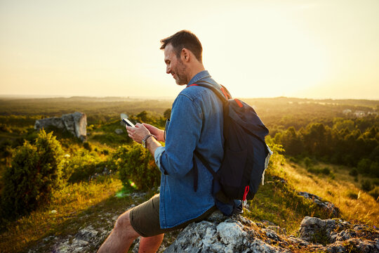 Adult man using phone hiking during sunset sitting on top of the mountain