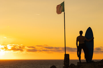 Silhouette of a man surfer standing by the ocean at sunset - the flag of Portugal on the flagpole