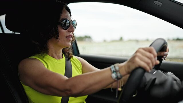 Middle age hispanic woman driving a car smiling wearing sunglasses on the road