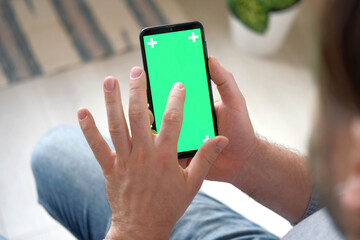 Young man sitting at home holding smartphone green mock-up screen in hand. Male person using chroma...