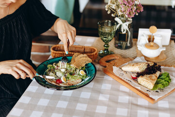 Tasting caesar salad using cutlery in female hands, which is on a table with a checkered beige tablecloth on it, next to which is a cheese pita on a wooden board, a wooden stand for salt, pepper