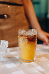 A mix of orange juice, coffee and ice in a decorative glass standing on a beige checkered tablecloth on a table against the background of a barista in a brown apron